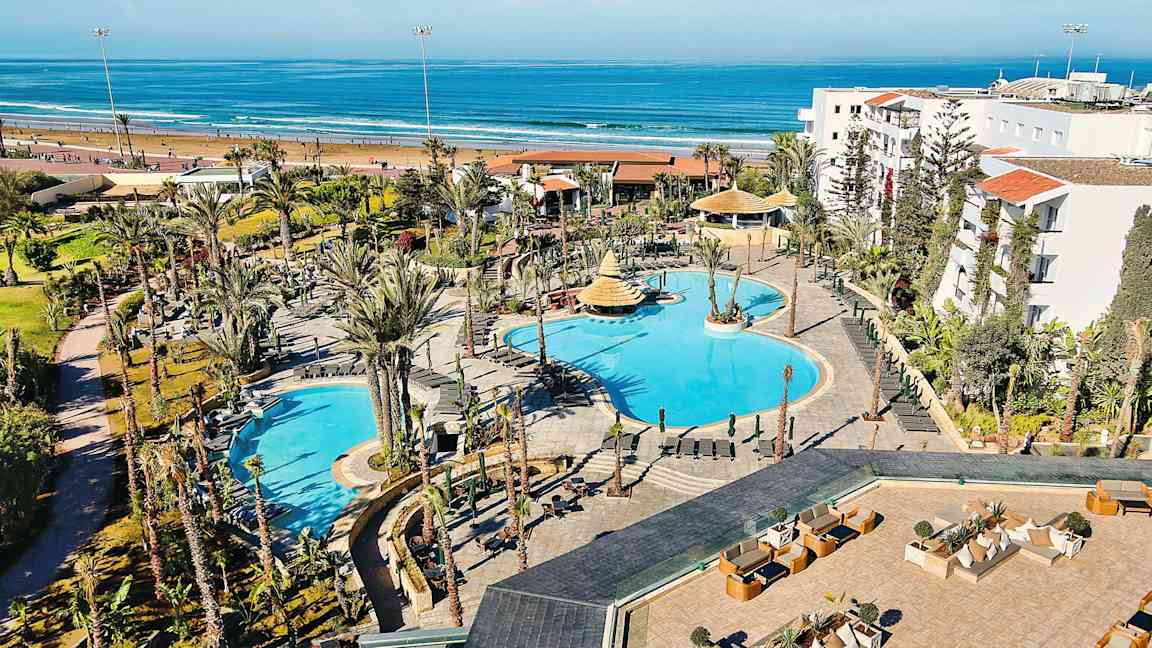 Hotelområde med pools og omkringliggende haver, beliggende tæt på en sandstrand og havet. (AI-genereret tekst)