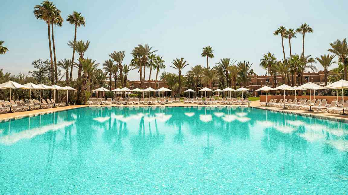 En stor pool omgivet af liggestole og parasoller under en klar himmel med palmer i baggrunden. (AI-genereret tekst)