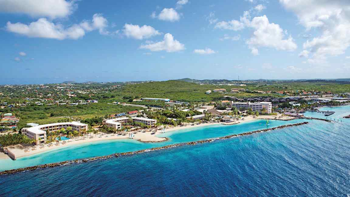 Luftfoto af en kystlinje med hotelbygninger ved en strand og grønne områder i baggrunden. (AI-genereret tekst)