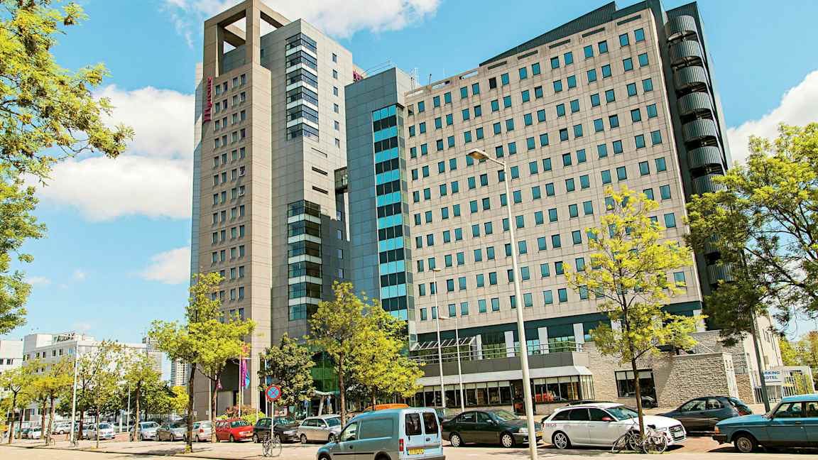 En moderne bygning med flere etager omgivet af parkerede biler og træer. (AI-genereret tekst)