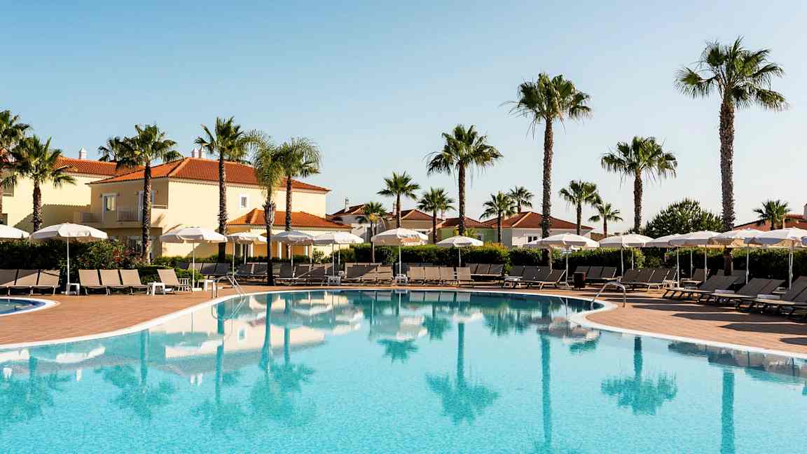 Udendørs swimmingpool med liggestole og parasoller, omgivet af palmer og bygninger med terrakottatag. (AI-genereret tekst)