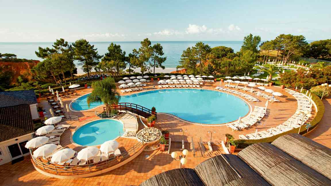 Udsigt over en swimmingpool med liggestole og parasoller, omgivet af grønne områder og med havet i baggrunden. (AI-genereret tekst)