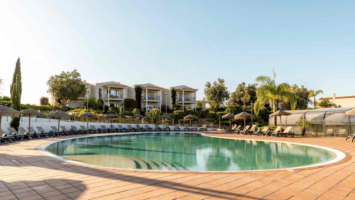 Udendørs pool med liggestole og parasoller, omgivet af grønt område og bygninger. (AI-genereret tekst)