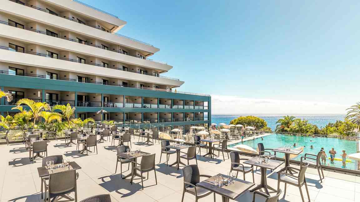 Udendørs serveringsområde med borde og stole foran et hotel og en pool med badende gæster, med havet i baggrunden. (AI-genereret tekst)