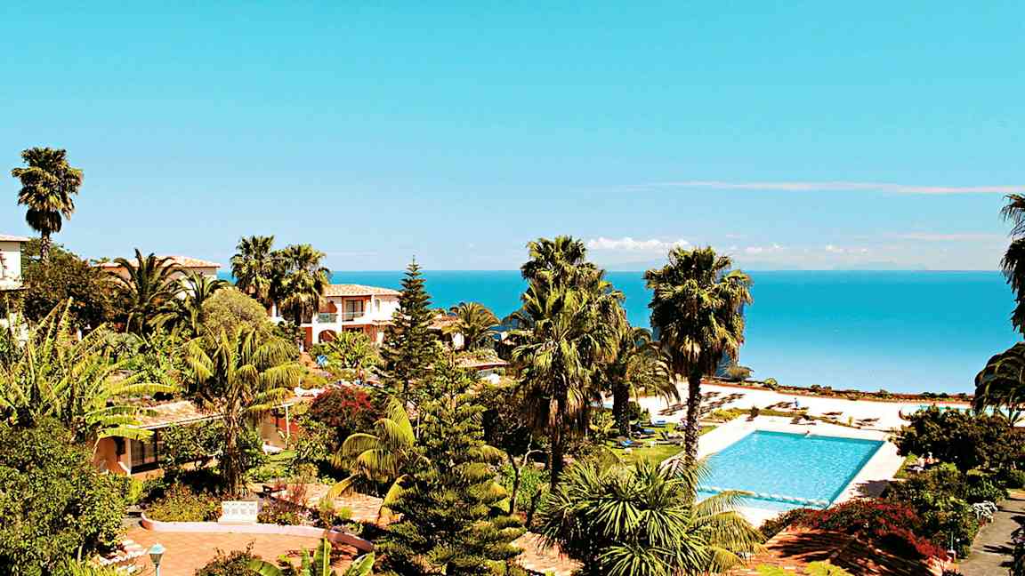 Udsigt over en tropisk have med palmer, pool og hav i baggrunden. (AI-genereret tekst)