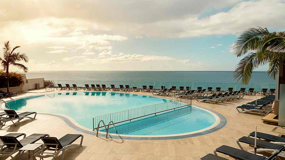 Udendørs pool med liggestole omkring, beliggende tæt på havet under en klar himmel. (AI-genereret tekst)