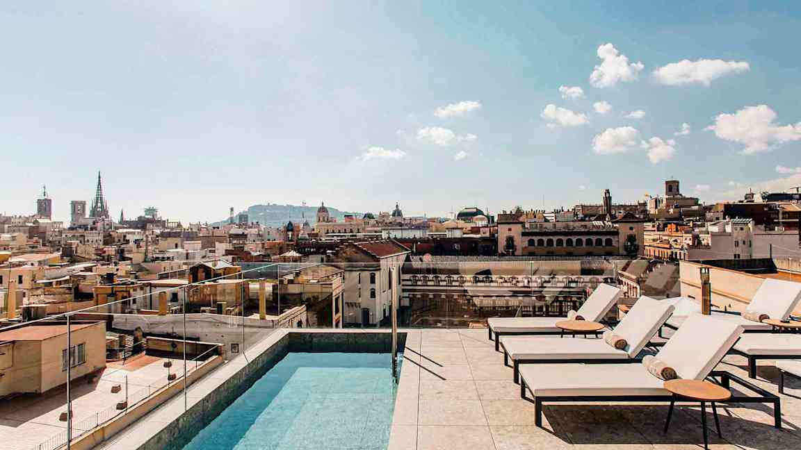 En udendørs pool og liggestole på en tagterrasse med udsigt over en by og dens bygninger under en klar himmel. (AI-genereret tekst)