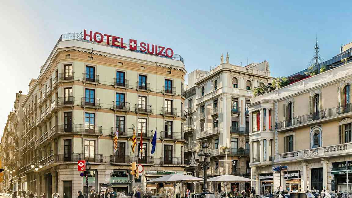 Et hotel ved navn "Hotel Suizo" på et hjørne med flere etager og altaner, omgivet af andre bygninger og folk, der går på gaden. (AI-genereret tekst)