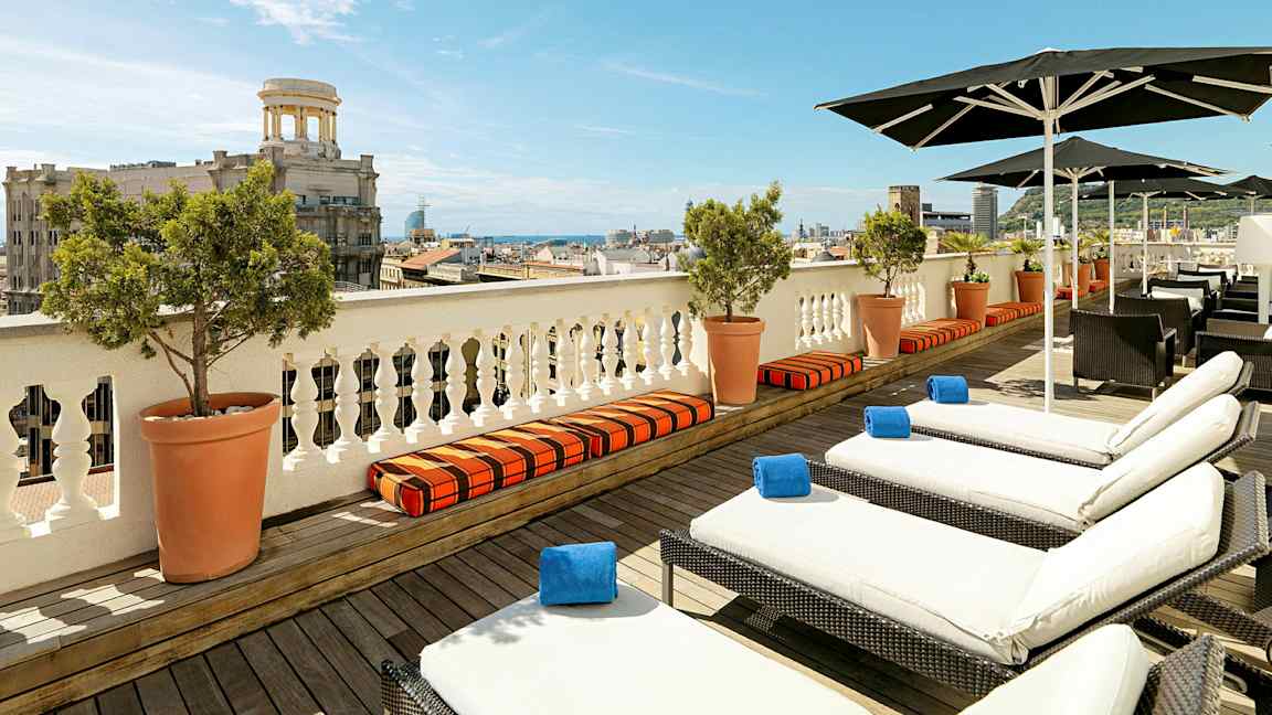 Udsigt over en terrasse med liggestole og parasoller, med en by og havet i baggrunden. (AI-genereret tekst)