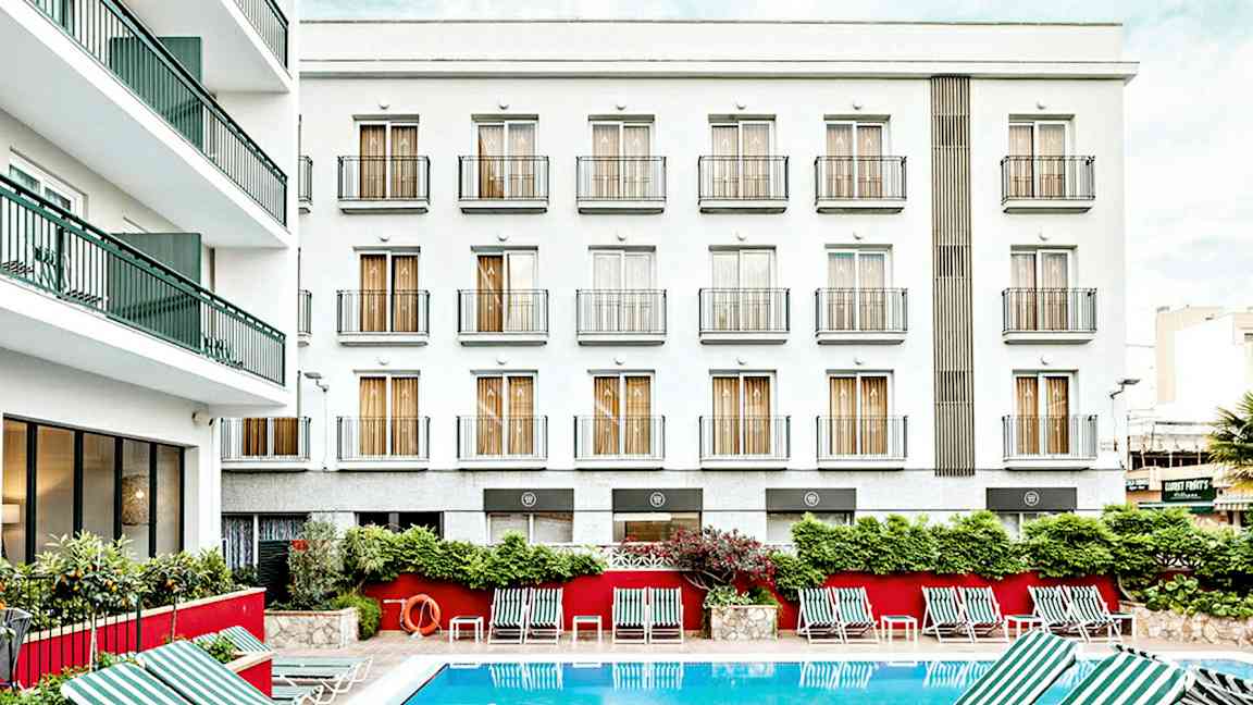 Billede af en hotelbygning med hvid facade og flere altaner. Foran bygningen er en udendørs pool med stribede liggestole placeret omkring. (AI-genereret tekst)