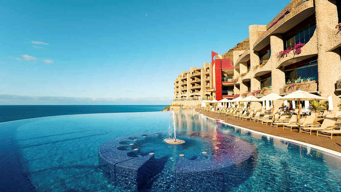 En infinitypool ved strandkanten med solsenge og hotelbygninger i baggrunden. (AI-genereret tekst)
