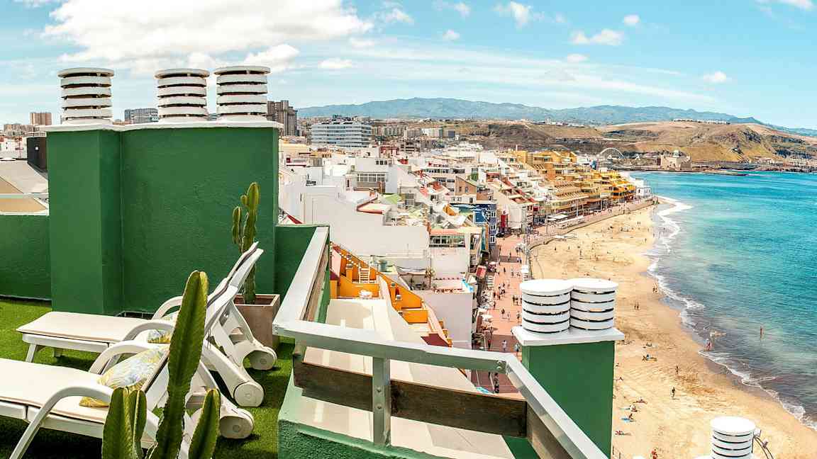 Udsigt fra en tagterrasse med solvogne, kaktusser og en strand samt byen i baggrunden. (AI-genereret tekst)