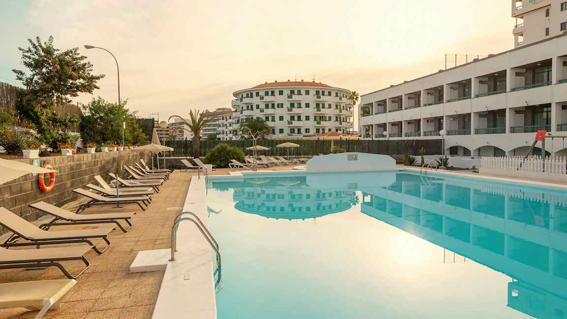 Udsigt over en pool med solsenge ved siden af og omkringliggende bygninger. (AI-genereret tekst)