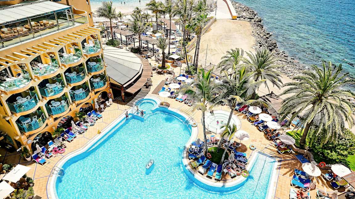 En hotelpool ved kysten med palmer og liggestole omkring. (AI-genereret tekst)