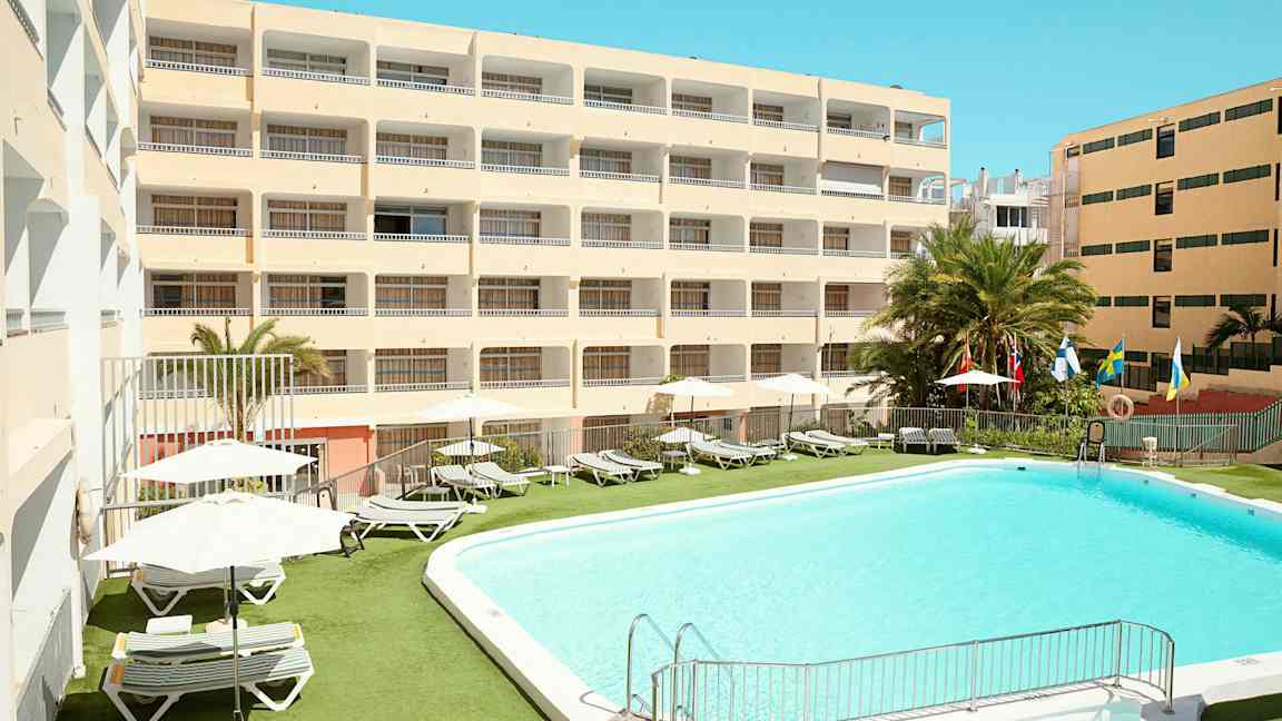 Hotelbygning med swimmingpool og liggestole udendørs. (AI-genereret tekst)