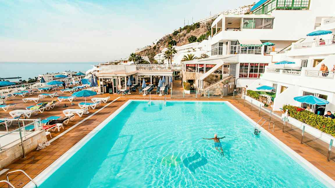 Billede af en stor udendørs pool omgivet af liggestole og parasoller med hvide bygninger og havet i baggrunden. (AI-genereret tekst)