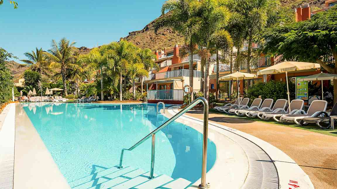 Udendørs pool med liggestole ved et hotel, omgivet af palmer og grønne planter. (AI-genereret tekst)