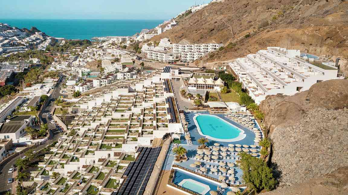 Luftfoto af et hotelområde med pools, liggestole og terrasser, beliggende ved en skråning nær kysten. (AI-genereret tekst)