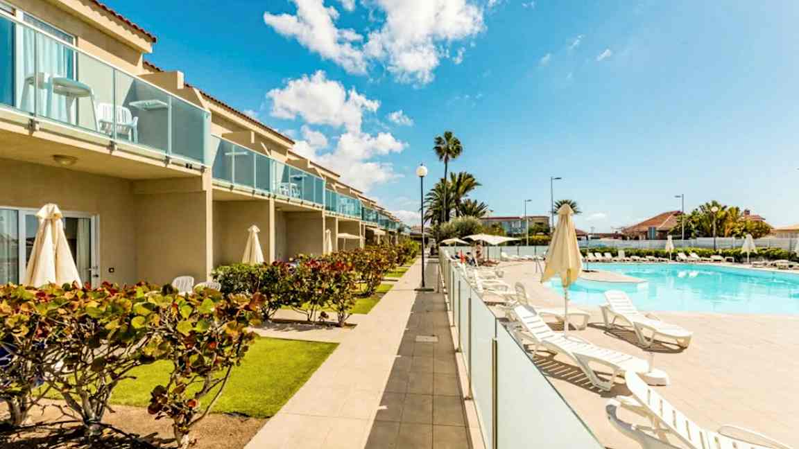 Billede af et hotelområde med pool, liggestole og grønne omgivelser. Bygningen har altaner. (AI-genereret tekst)