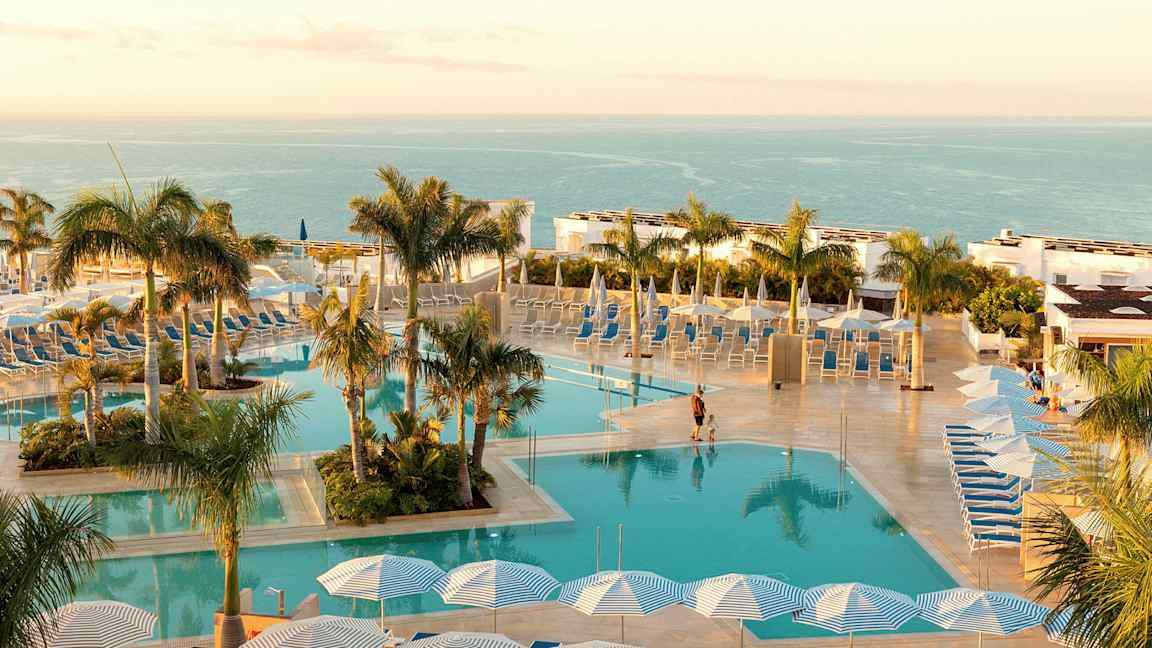 En pool med liggestole og parasoller, omgivet af palmer med havet i baggrunden. (AI-genereret tekst)