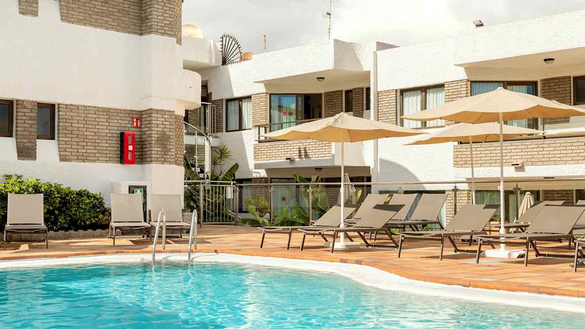 Udendørs poolområde med liggestole og parasoller ved en bygning. (AI-genereret tekst)