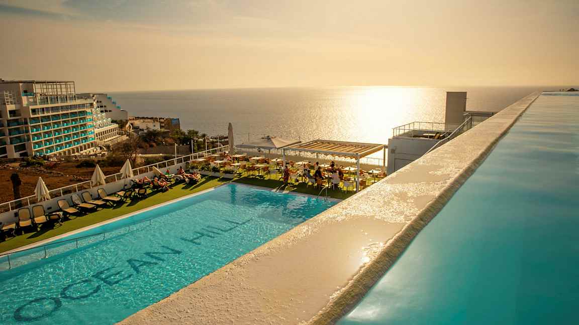 Udendørs swimmingpool med liggestole og udsigt over havet ved solnedgang. (AI-genereret tekst)