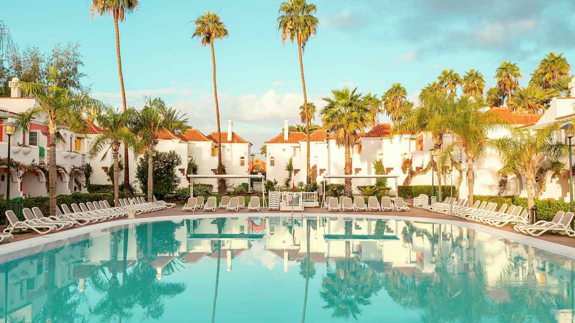 Hotelområde med hvidkalkede bygninger med røde tage, omgivet af palmer, og en stor pool med liggestole omkring. (AI-genereret tekst)