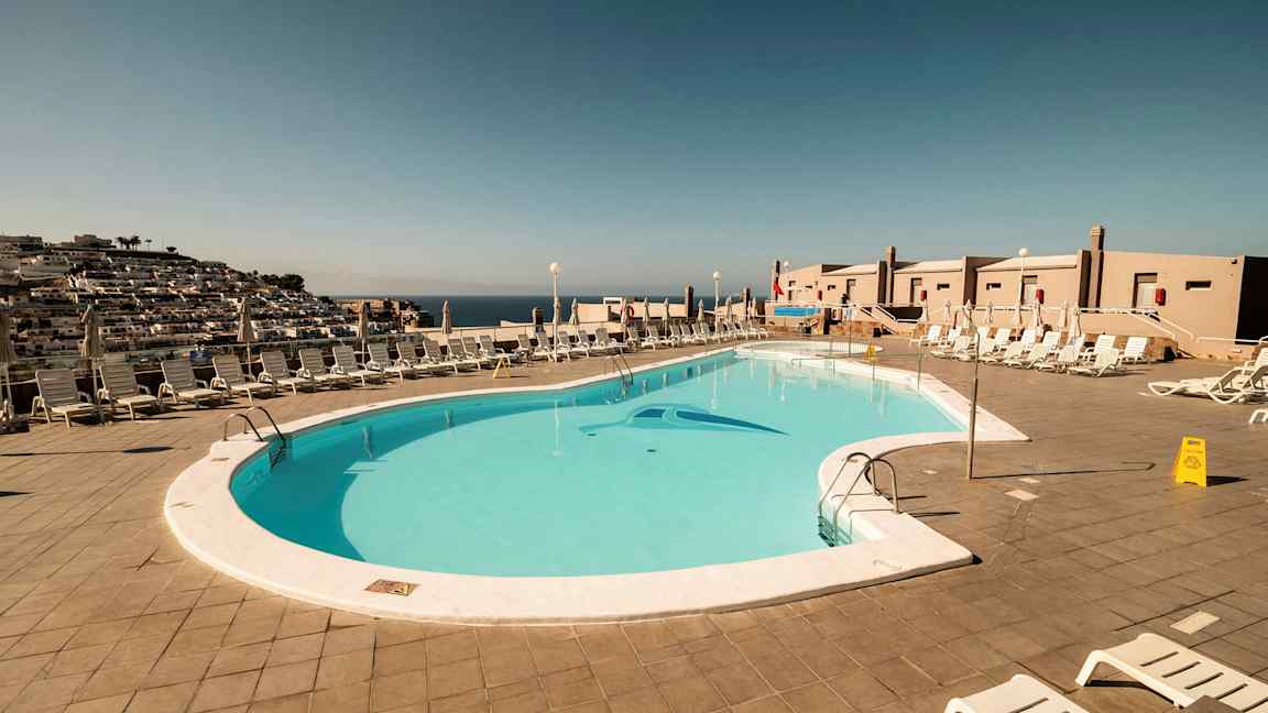 Udsigt over en udendørs swimmingpool omgivet af liggestole og parasoller. (AI-genereret tekst)