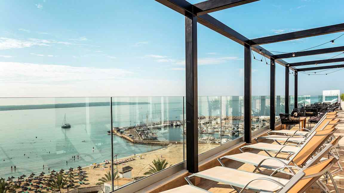 Liggestole opstillet foran glasværn med udsigt over strand og havn. (AI-genereret tekst)