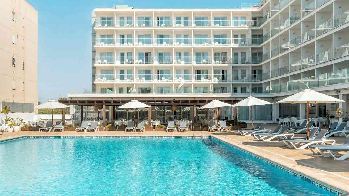 En bygning med flere altaner, foran en pool med liggestole og parasoller. (AI-genereret tekst)