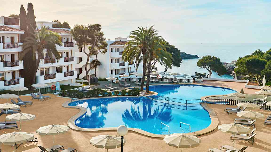 Billede af et hotelområde med en stor pool omgivet af liggestole og parasoller. I baggrunden ses havet og dele af bygningen. (AI-genereret tekst)