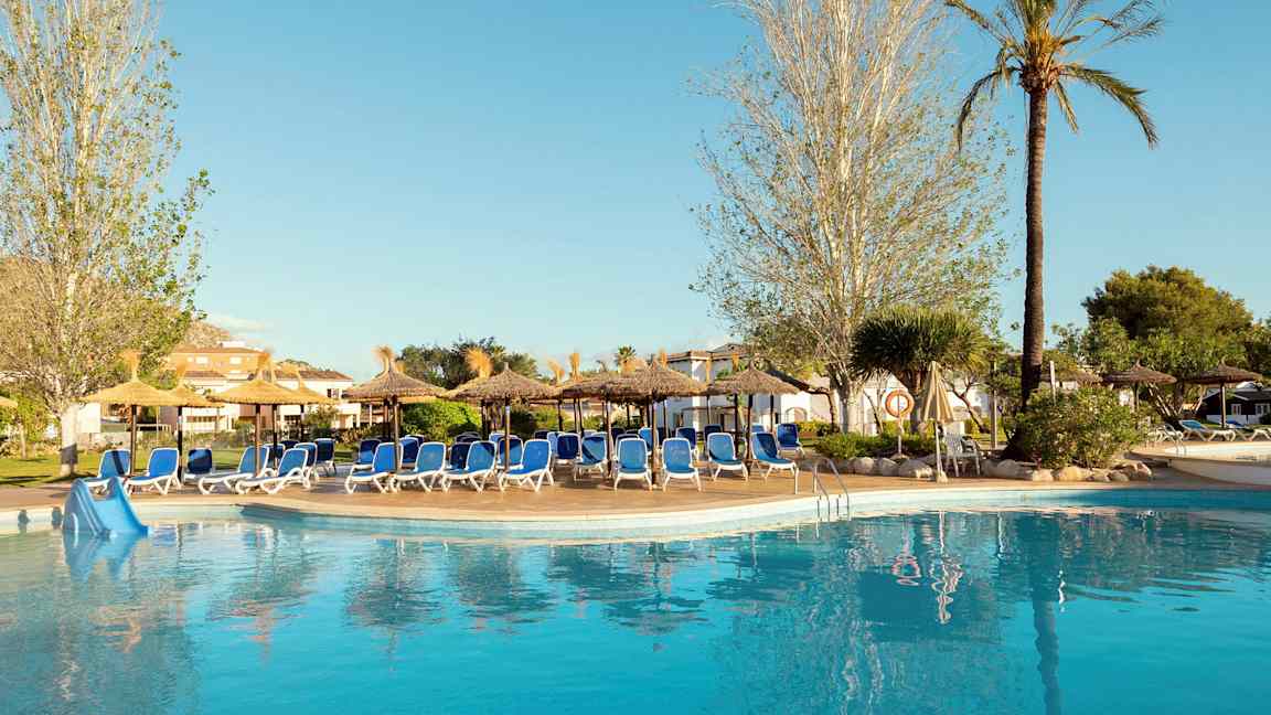 En pool ved et hotel omgivet af liggestole og parasoller, omgivet af træer og beplantning. (AI-genereret tekst)