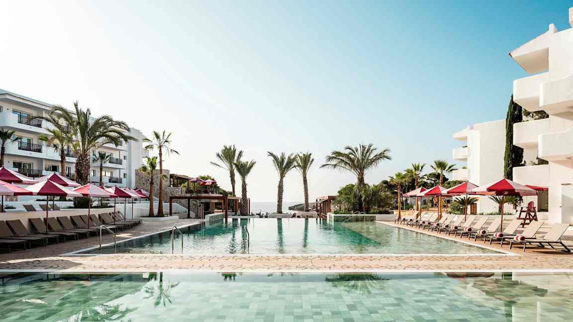 Udendørs pool ved kystnært hotel med liggestole og røde parasoller. (AI-genereret tekst)