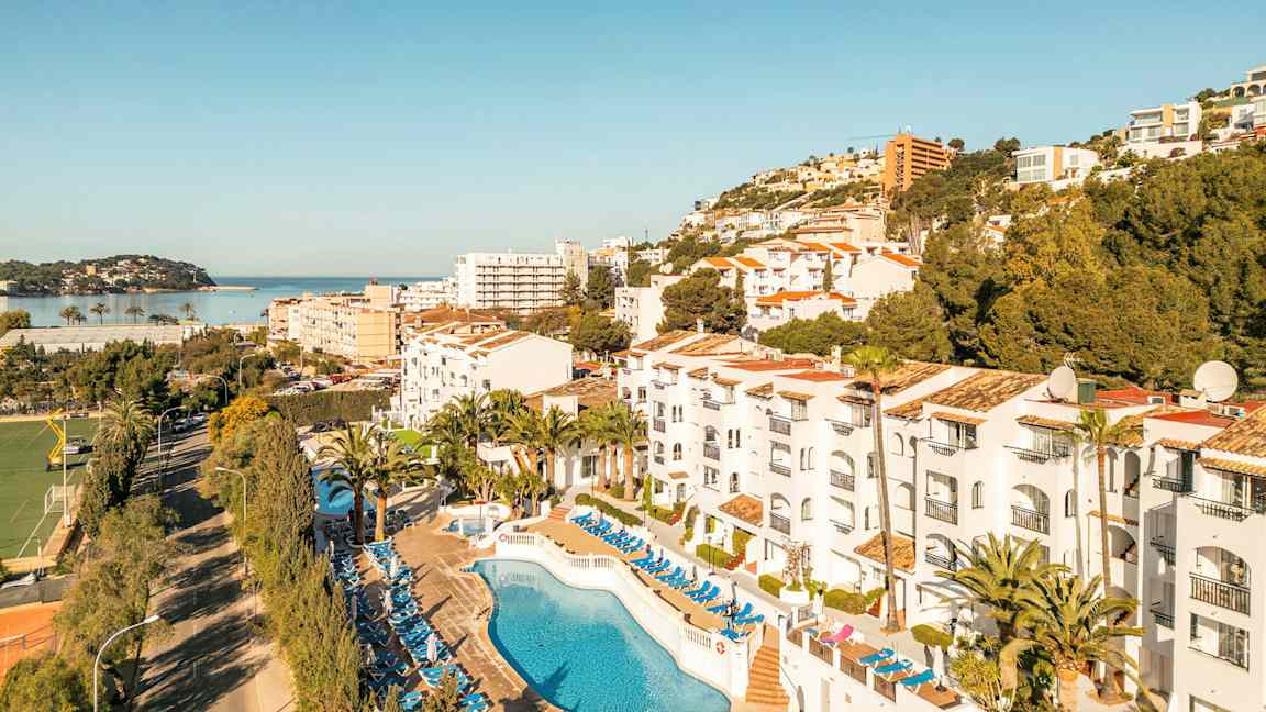 Billede af hotelområde med pool og liggestole, omgivelser med træer og bygninger samt udsigt mod havet. (AI-genereret tekst)