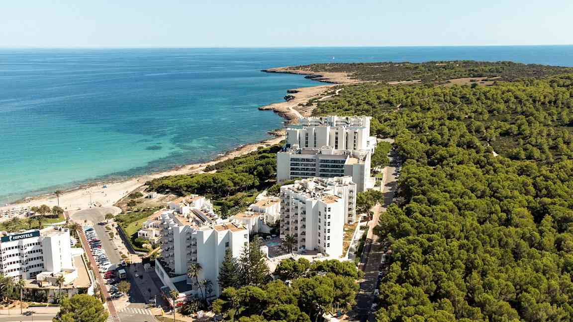 Luftfoto af en kystlinje med hotelbygninger foran en skov og tæt på en strand. (AI-genereret tekst)