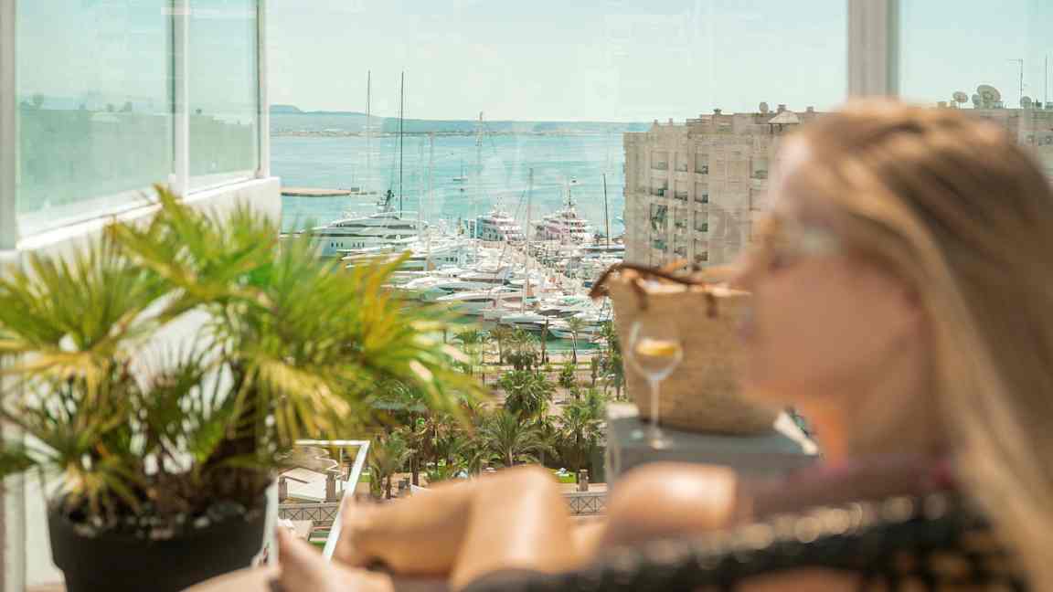 Udsigt mod både i en marina, med en glasaltan og frodige planter i forgrunden. (AI-genereret tekst)