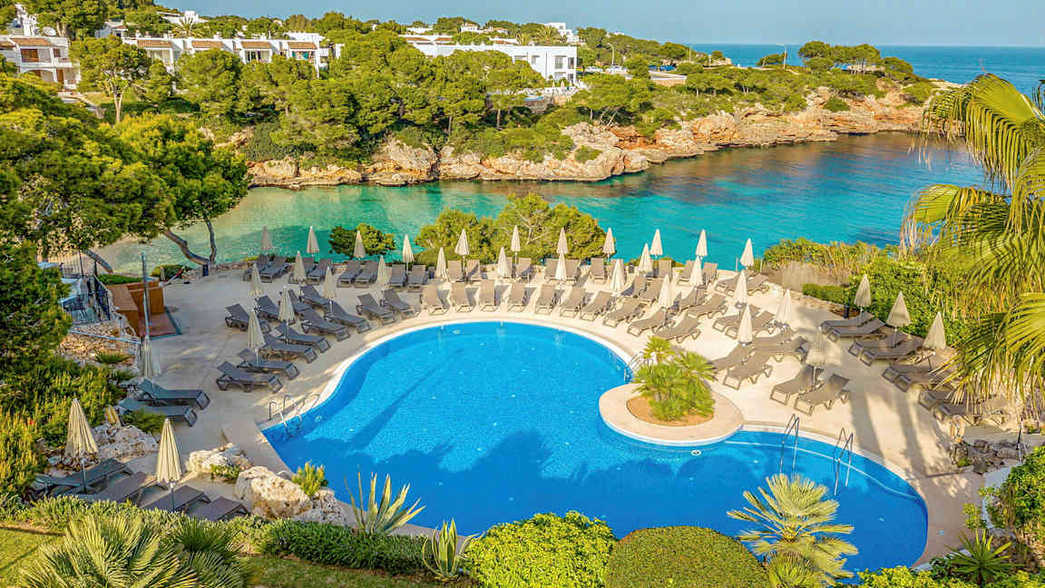 En udendørs pool med liggestole og parasoller, omgivet af grønt og med udsigt til havet. (AI-genereret tekst)