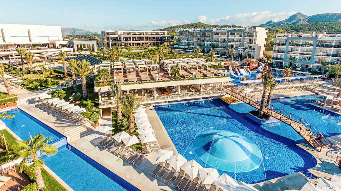 En udsigt over et hotelområde med pools, liggestole og bygninger. (AI-genereret tekst)