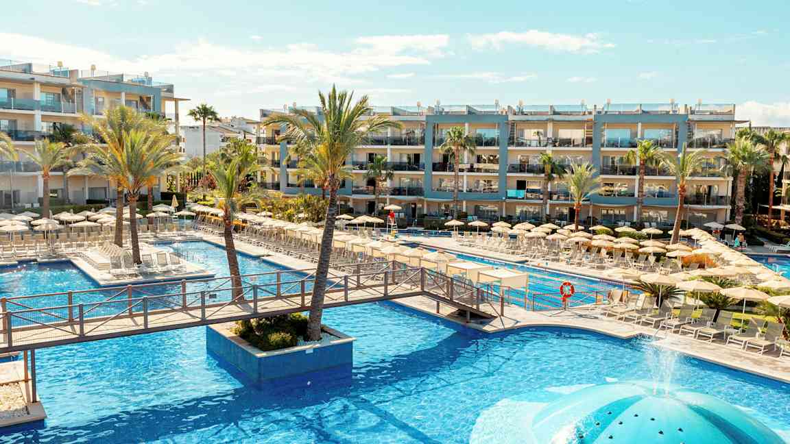 Poolområde ved et hotel med liggestole og parasoller, omgivet af palmer og bygninger. (AI-genereret tekst)