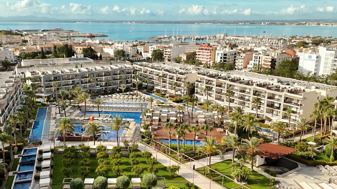 Et hotelkompleks med pools omgivet af grønne områder og liggestole, beliggende ved vandet med en havn i baggrunden. (AI-genereret tekst)