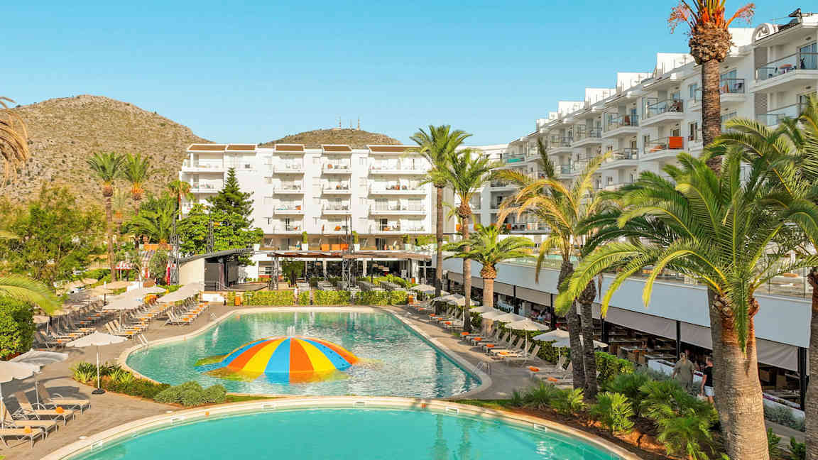 Et hotel med hvide bygninger, liggestole og en pool med en farverig vandstrålende kuppel i midten. (AI-genereret tekst)