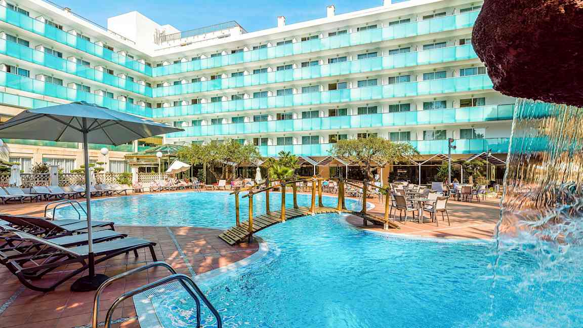 En udendørs swimmingpool ved en hotelbygning med liggestole, borde og parasoller. En lille bro over poolen og en del af en vandfaldsstruktur ses. (AI-genereret tekst)