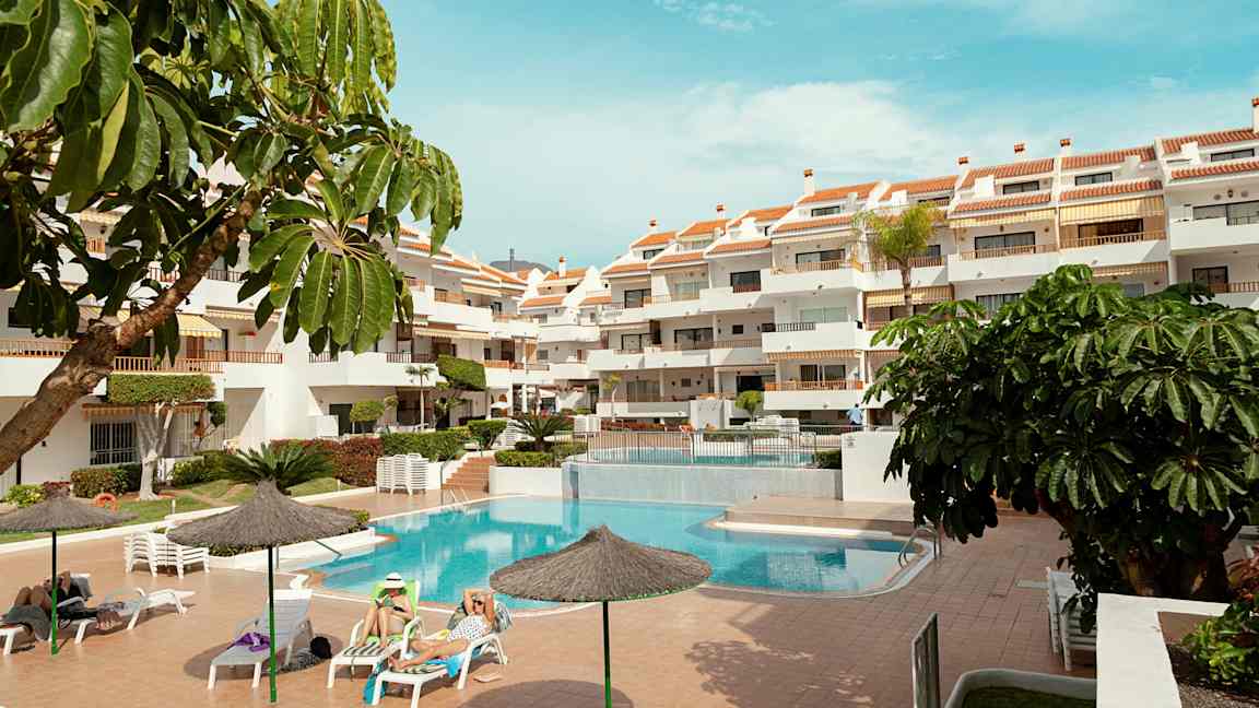 Et udendørs område med en pool, liggestole og et fleretagers hotel. Flere parasoller og grønne planter omgiver området. (AI-genereret tekst)