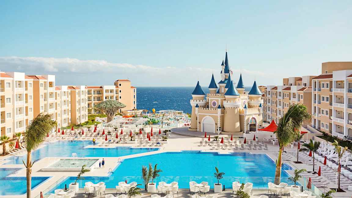 Et feriested med swimmingpools, en bygning der ligner et eventyrslot og omkringliggende hotelbygninger nær havet. (AI-genereret tekst)