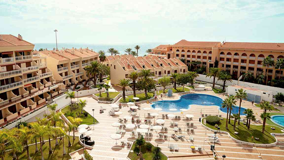 Flere bygninger med terrakottatage omkranser en pool og solsenge, med omkringliggende palmer og havet i baggrunden. (AI-genereret tekst)