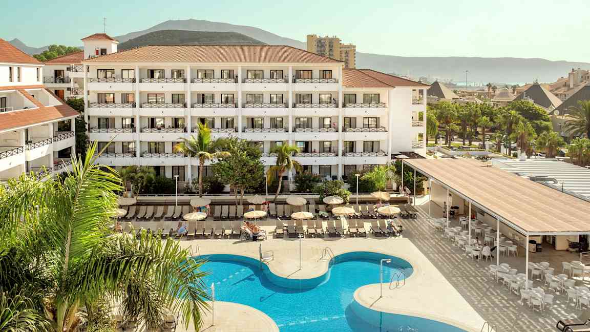 Hotelkompleks med pool og liggestole, omgivet af grønne områder og bjerge i baggrunden. (AI-genereret tekst)