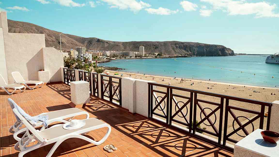 Terrasse med liggestole og udsigt over en sandstrand med havet i horisonten. (AI-genereret tekst)
