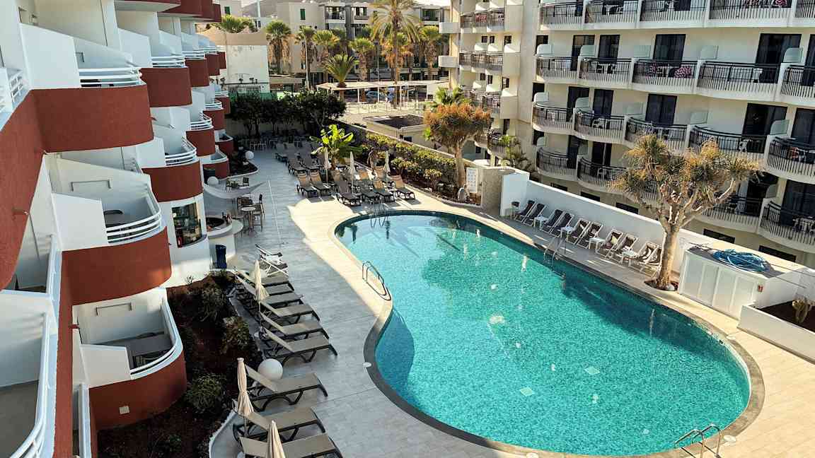 Et hotel med flere altaner og en formet pool omgivet af liggestole og parasoller. (AI-genereret tekst)