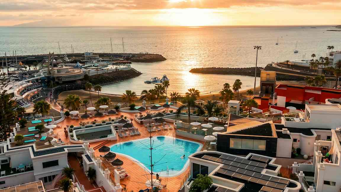 Udsigt over et resort med swimmingpools, liggestole og parasoller, med en strand og lystbådehavn ved kysten. Solnedgangens lys reflekteres i vandet. (AI-genereret tekst)