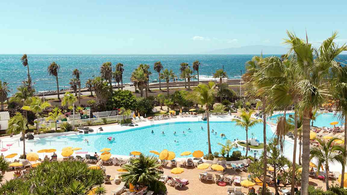 Poolområde med liggestole, parasoller og mennesker, med havet i baggrunden. (AI-genereret tekst)